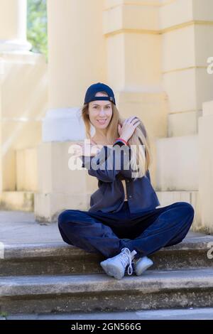 Lächelnde langhaarige blonde Frau in Sportkleidung sitzt auf einer Treppe im Park Stockfoto