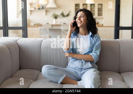 Glückliche junge kaukasische Dame, die auf der Couch sitzt, zu Hause mit dem Mobiltelefon telefoniert, freier Platz Stockfoto