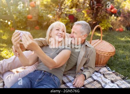 Moderne ältere Ehepartner, die Selfie auf dem Smartphone machen, Picknick machen und sich am sonnigen Herbsttag im Garten ausruhen Stockfoto