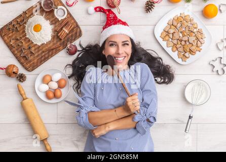 Glückliche dunkelhaarige Frau in weihnachtshut mit einem Holzlöffel und lag auf dem Boden mit traditionellen Zutaten wie Mehl, Eier, Orangen und al Stockfoto