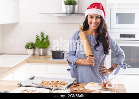 Weihnachtsmutter trägt einen weihnachtshut und hält die Rolle glücklich, während sie Christkindgebäck in ihrer Küche backt. Stockfoto