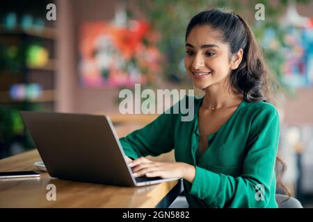 Hübsche indische Frau, die online im Café studiert, Laptop benutzt, leeren Raum Stockfoto