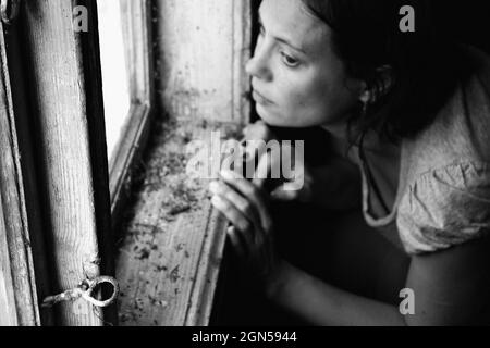 Surreales Porträt. Frau schaut durch das alte Fenster. Porträt einer geheimnisvollen jungen Frau. Schwarzweiß-Fotografie. Stockfoto