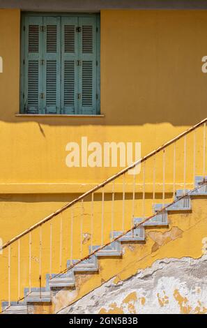 Ein Fenster mit Fensterläden, das grün in einer hellgelben Wand mit einer Steintreppe oder einer Treppe in einem typisch griechischen Gebäude gestrichen wurde. Flug aus Steintreppen. Stockfoto