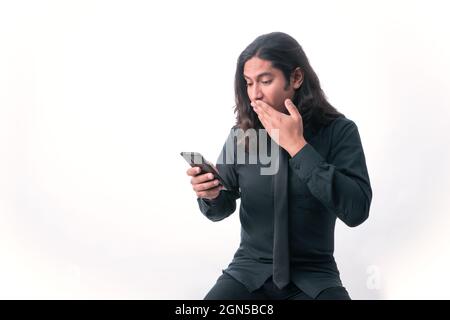 Person mit weißem Hintergrund. Er schaut auf sein Handy, hält seine Hand überrascht über etwas, das er sich angeschaut hat. Er ist angezogen Stockfoto