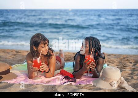 Unbeschwerte junge multirassische Freundinnen, die auf einer Decke liegen und erfrischende rote Getränke genießen, während sie am Sommertag am Sandstrand gemeinsam chillen Stockfoto