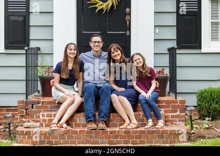 Eine Familie mit einer Mutter, einem Vater und zwei Töchtern, die draußen auf der Backsteinvertei eines kleinen blauen Häuschens sitzen Stockfoto