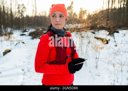 Weibliche Läuferin hält mobil auf der Suche nach einem Weg aus dem Wald im Winter. Sportlerin schaut sich im Wald um und überprüft die Position auf dem Mobiltelefon auf dem kalten Winter D Stockfoto