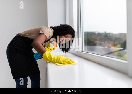 Portrait des jungen Afro-amerikanische Frau im gelben Handschuhe Fensterbänke Waschen mit Tuch und Glasreiniger im Innenbereich Stockfoto