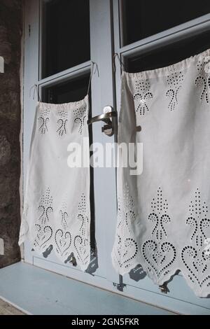 Blaues Fenster mit weißen Vorhängen mit schönem Stoff, der nachts im Innenbereich des Hauses in Herzmustern in Nahaufnahme dekoriert ist. Traditionelles mediterranes Haus. Version Stockfoto