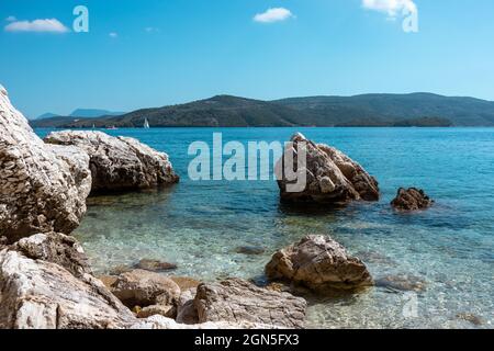 Große Felsen im türkisfarbenen Wasser an der Küste mit weißer Segelyacht und grünen Hügeln am Horizont, Insel Lefkada in Griechenland. Sommer lebendige Natur Reise nach Ionian Stockfoto