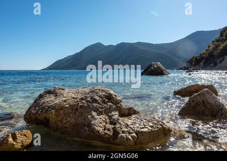 Große Felsen in kristallklarem azurblauem Wasser am sonnigen Kiesstrand mit grünen Hügeln am Horizont. Küste der Insel Lefkada in Griechenland. Sommer wilde Natur reisen Stockfoto