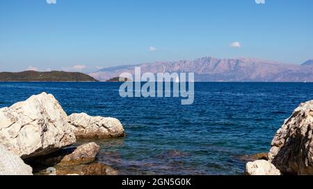 Große Felsen in blauem Wasser an der Küste mit weißer Segelyacht und Bergen am Horizont, Insel Lefkada in Griechenland. Lebendige, wilde Natur reisen zum Ionischen Meer Stockfoto