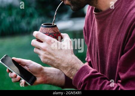 Mann trinkt einen Mate und hält ein Handy in der Hand. Mate Tee aus Südamerika. Stockfoto