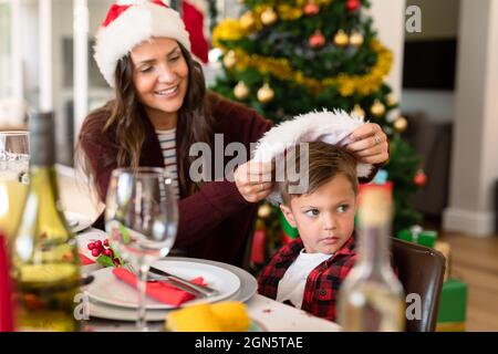 Kaukasische Mutter, die ihren Sohn am weihnachtstisch mit Weihnachtsmütze auf den Kopf setzt Stockfoto