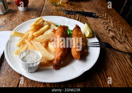 Eine Portion Fisch und Chips mit Erbsen und Tartarsauce auf einem weißen Porzellanplatte aus Keramik auf einem Tisch in einem Pub; traditionelle englische Küche. Stockfoto