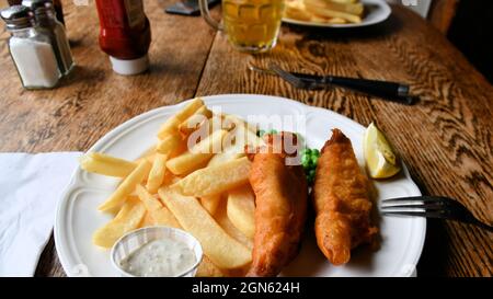 Eine Portion Fisch und Chips mit Erbsen und Tartarsauce auf einem weißen Porzellanplatte aus Keramik auf einem Tisch in einem Pub; traditionelle englische Küche. Stockfoto