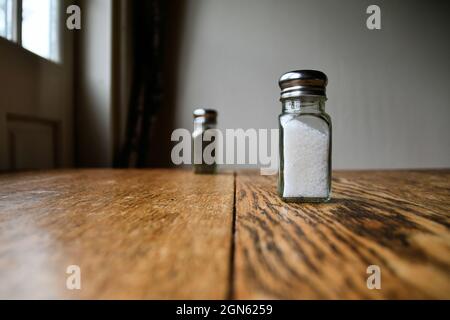 Salz- und Pfefferstreuer auf einer polierten Holzoberfläche mit Licht aus einem Fenster, das den Tisch beleuchtet, gemahlen; rustikaler Esstisch mit Glasstreuern. Stockfoto