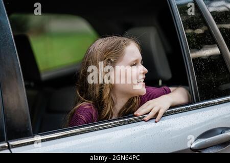 Kleines kaukasisches Mädchen mit Sommersprossen und einem lila Hemd, das aus einem Autofenster schaut Stockfoto