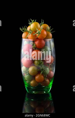 Kirschtomaten, rote, gelbe und grüne kleine runde Tomaten in einem Glas auf einer reflektierenden Oberfläche, isoliert auf schwarzem Hintergrund Stockfoto