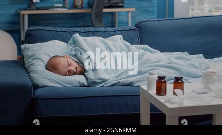Unwohl Erwachsener, der auf dem Sofa lag und mit Medikamenten auf dem Tisch schlief. Kranker Mann, der sich wegen Erkältung und Grippe ausruhte, mit Pillen, Medikamenten, Geweben und einer Tasse Tee. Person mit Krankheit Stockfoto