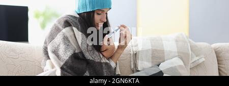 Junge Frau in warme Decke gewickelt sitzt auf dem Sofa mit Thermometer Stockfoto