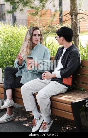Vertikale Vollaufnahme von zwei stylischen jungen Frauen in lässigen Outfits, die im Freien auf der Bank sitzen und Kaffee trinken und über etwas reden Stockfoto