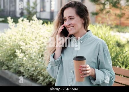 Horizontale Taille nach oben Porträtaufnahme der schönen jungen erwachsenen kaukasischen Frau mit langen welligen Haaren lächelnd, während Sie am Telefon sprechen und Kaffee im Freien trinken Stockfoto