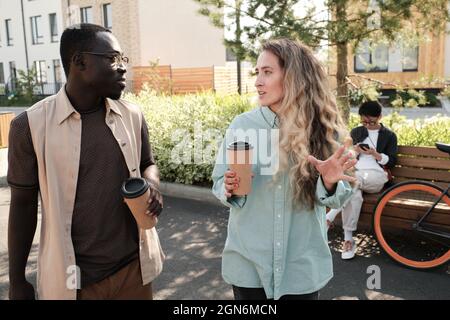 Horizontale, mittlere Porträtaufnahme eines jungen afroamerikanischen Mannes und einer kaukasischen Frau, die im Freien spazieren und Kaffee trinken und zwanglose Gespräche führen Stockfoto