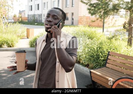 Horizontales mittleres Porträt eines hübschen jungen schwarzen Mannes, der im Freien spazieren geht, Kaffee trinkt und mit jemandem telefoniert Stockfoto