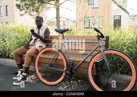 Horizontale Langaufnahme eines modernen jungen afroamerikanischen Mannes, der ein legeres Outfit trägt und entspannt auf einer Holzbank sitzt und etwas auf dem Smartphone schreibt Stockfoto
