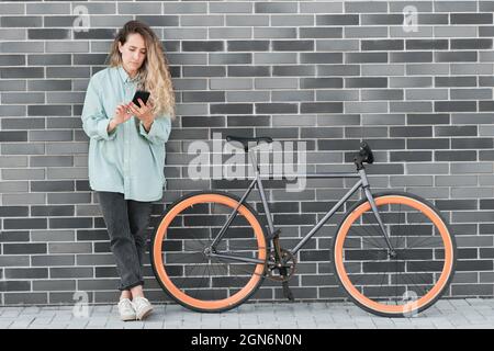 Junge kaukasische Frau, die sich im Freien an einer grauen Backsteinmauer lehnt, mit einem Fahrrad, das neben ihr geparkt ist und auf dem Smartphone etwas im Internet beobachtet Stockfoto