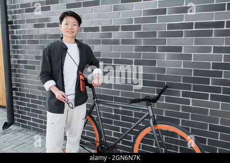Horizontale, mittellange Aufnahme einer jungen asiatischen Frau mit kurzen Haaren in modischem Freizeitoutfit, die mit dem Fahrrad gegen ein graues Backsteingebäude steht und die Kamera anlächelt Stockfoto