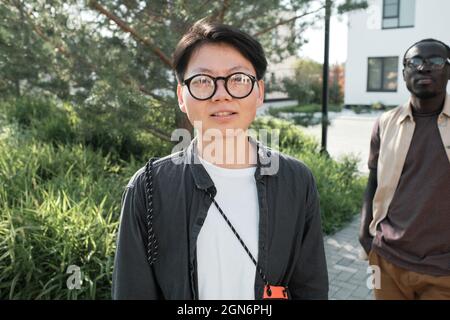 Horizontales mittleres Porträt einer schönen jungen asiatischen Frau mit Hemdhaarschnitt in einem lässigen Outfit mit einer Brille, die im Freien steht und auf die Kamera schaut Stockfoto