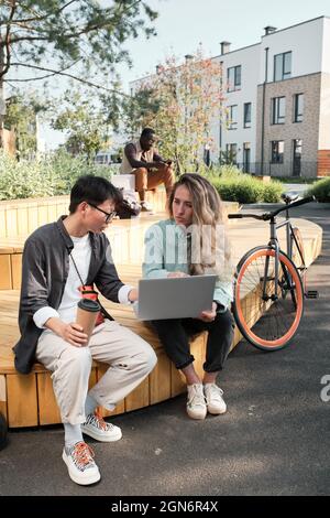Vertikale Aufnahme von zwei jungen Kollegen, die im modernen Park im Freien sitzen und sich etwas auf dem Laptop anguckend über Probleme unterhalten Stockfoto