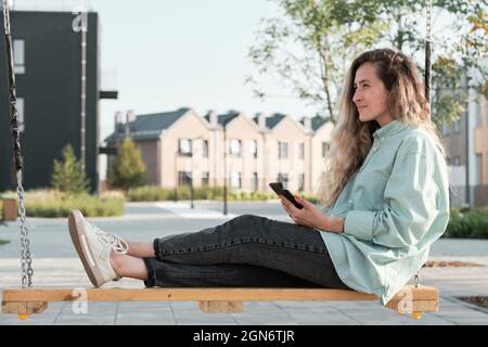 Horizontale Seitenansicht Aufnahme einer schönen jungen erwachsenen kaukasischen Frau mit welligen Haaren, die entspannt auf der Schaukel sitzt und das Smartphone an etwas denkt Stockfoto