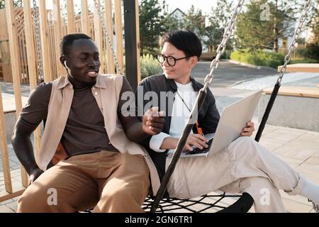 Horizontales mittleres Porträt eines modernen afroamerikanischen Mannes und einer asiatischen Frau, die auf Schaukel im Park sitzen und an einem Projekt arbeiten Stockfoto