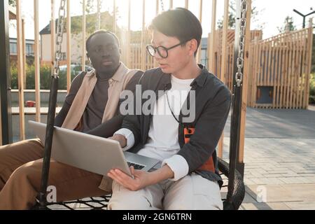 Horizontales mittleres Porträt eines schönen afroamerikanischen Mannes und einer stilvollen asiatischen Frau, die im Stadtpark zusammen auf Schaukel sitzen und an einem wichtigen Projekt arbeiten Stockfoto