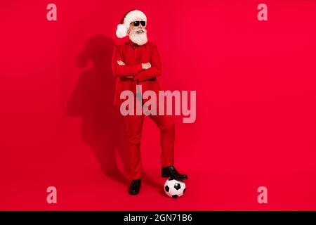 Foto von lustigen coolen Mann gekleidet weihnachten Kostüm Hut Arme gefaltet spielen Fußball leeren Raum isoliert roten Hintergrund Stockfoto
