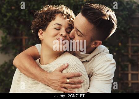 Flirtiger junger Mann, der seine Freundin im Freien küsst. Romantischer junger Mann, der seine Freundin auf die Wange küsst und sie von hinten umarmt. Junge LGBTQ+ Stockfoto