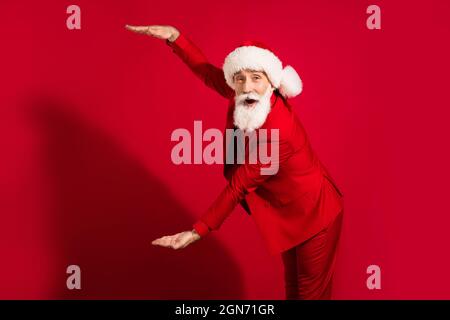 Foto von beeindruckt weißen Haaren gealterten Mann Hände messen tragen santa Kappe roten Anzug auf rotem Hintergrund isoliert Stockfoto