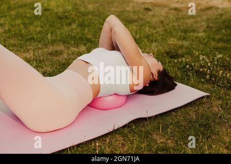 Nahaufnahme einer schlanken Frau in rosa oben, die Arme um die Schultern umarmt und mit Schulterblättern auf einem kleinen Gummiball im Sommer im Park liegt, Konzept von Stockfoto