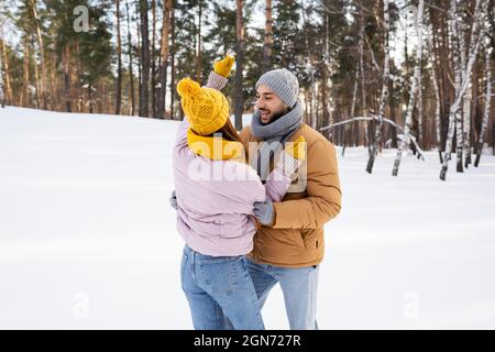 Lächelnder Mann, der im Park mit Schnee die Freundin in einem gestrickten Hut ansieht Stockfoto