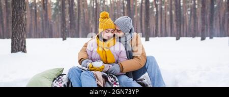 Junger Mann küsst Freundin im Winter Outfit mit Tasse im verschneiten Park, Banner Stockfoto
