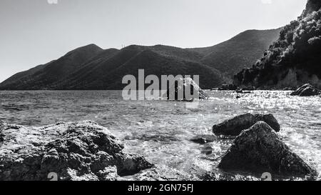 Schwarze und weiße große Felsen in kristallklarem Wasser am sonnigen Kiesstrand mit Hügeln am Horizont. Küste der Insel Lefkada in Griechenland. Sommer wilde Natur tr Stockfoto