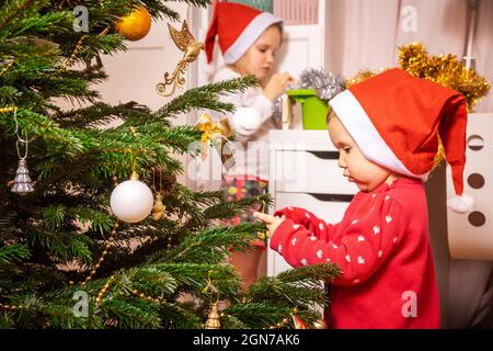 Kleines Mädchen trägt roten weihnachtspullover und weihnachtsmann Hut hilft, Weihnachtsbaum zu Hause zu schmücken Stockfoto