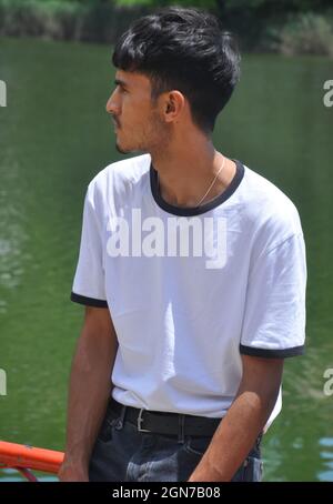 Ein gut aussehender indischer junger Mann, der weißes T-Shirt und schwarze Jeans trägt und auf einer Sicherheitsbarriere am See sitzt und zur Seite schaut Stockfoto