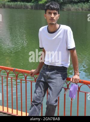 Ein hübscher junger Mann aus dem Norden Indiens, der weißes T-Shirt und schwarze Jeans trägt und eine Gesichtsmaske an der Hand hält, auf einer Sicherheitsbarriere am See sitzt und die Kamera anschaut Stockfoto