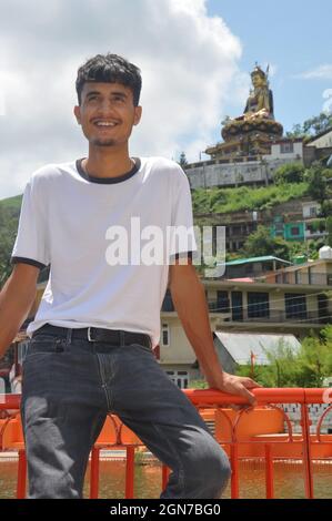 Ein junger Mann, der lächelt und auf einer Sicherheitsbarriere neben dem See sitzt, mit Blick weg und einer wunderschönen hügeligen Gegend von Tso-Pema und einer Statue von Padmasambhava (Guru Rinpoche) im Hintergrund Stockfoto