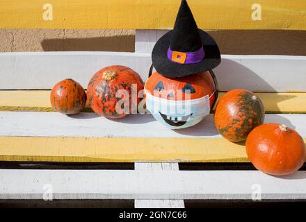 Fünf leuchtend orange Kürbisse in Reihe auf Holzbank. Einer von Kürbissen in Hexenhut und medizinischer Maske mit bemaltem gruseligen Lächeln. Straßendekorationen für Hal Stockfoto